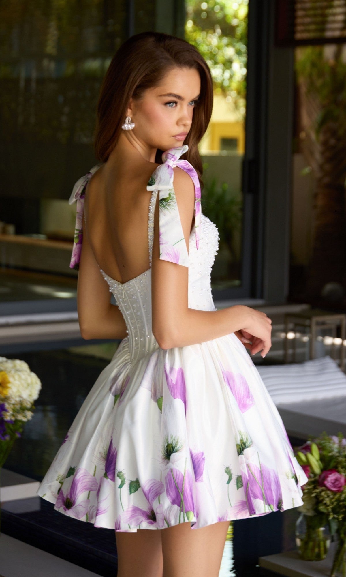 A woman in the Ava Presley 42432 short cocktail dress with floral print satin and bow straps stands indoors, looking over her shoulder.